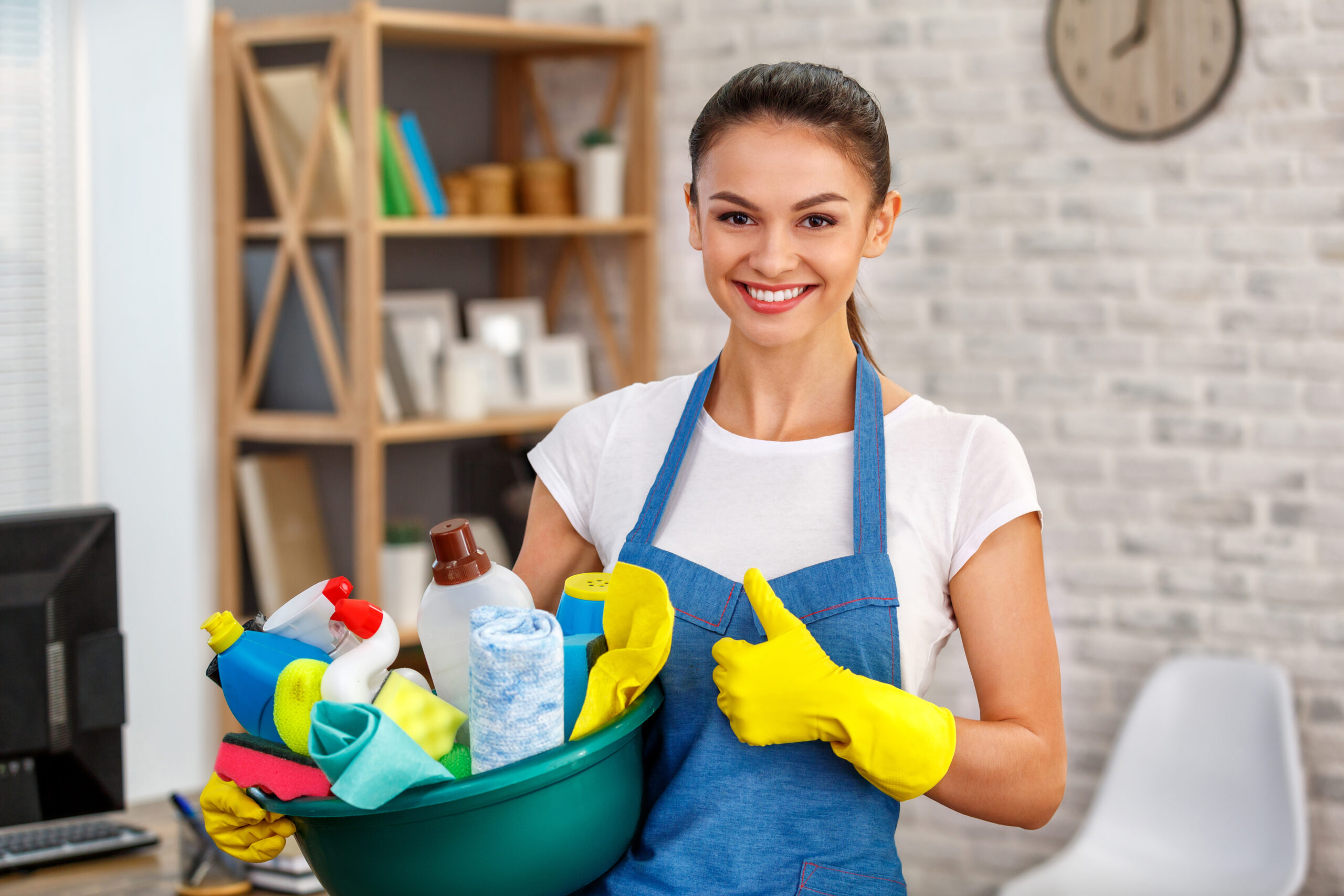 Studio shot of housekeeper. Beautiful woman cleaning office. Woman wearing gloves, smiling, showing thumb up and holding bowl full of bottles with disinfectant